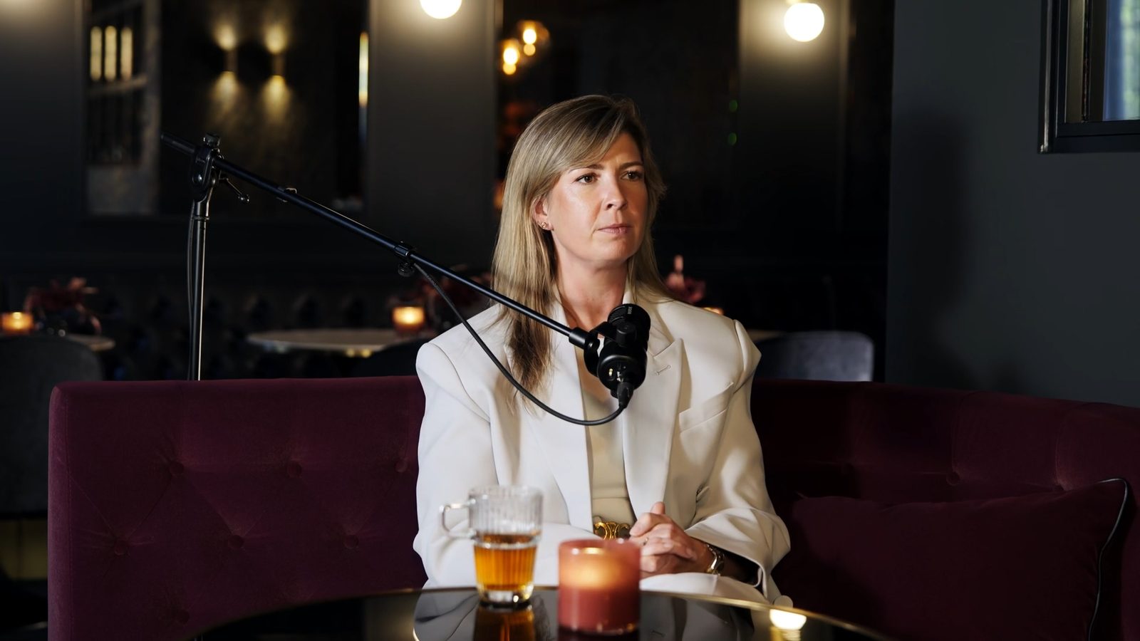 Podcastgast achter een microfoon aan de bar van een luxe hotelvergaderruimte, met drankjes op tafel.