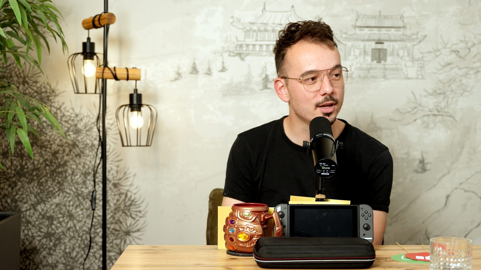 Podcastgast achter een microfoon aan tafel thuis, met een spelcomputer en een grote muurtekening als backdrop.