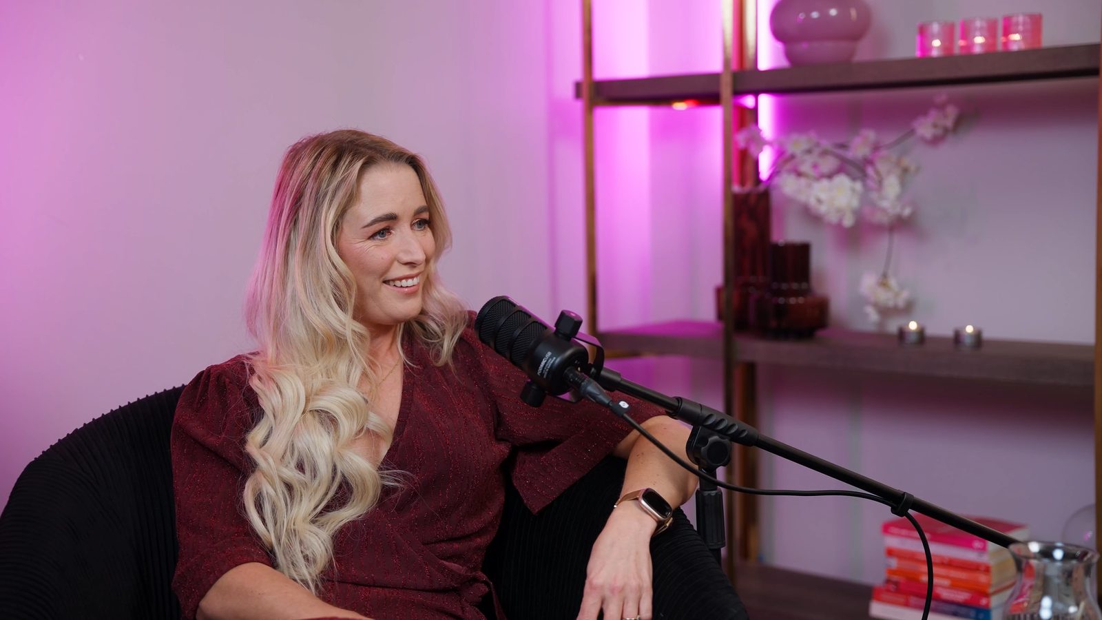 Podcastgast achter een microfoon in een interieur dat is uitgelicht in roze merkkleuren, met kaarsen en boeken.